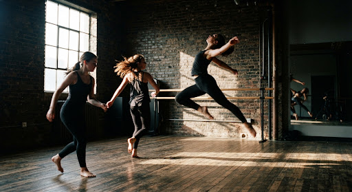 Intense high-contrast shot of dance students rehearsing in an atmospheric loft dance studio with natural sunlight streaming across wooden floorboards and brick walls